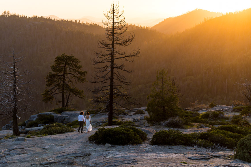 A bride and groom standing on a rocky cliff at sunset.