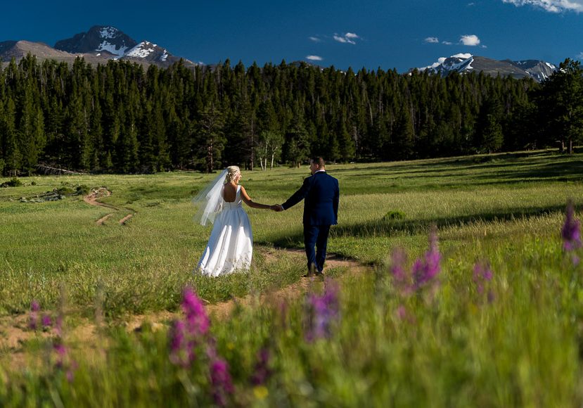 A bride and groom walking through a meadow with mountains in the background. A bride and groom walking through a meadow with mountains in the background.