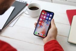 A man holding an iphone x on a desk with a cup of coffee.