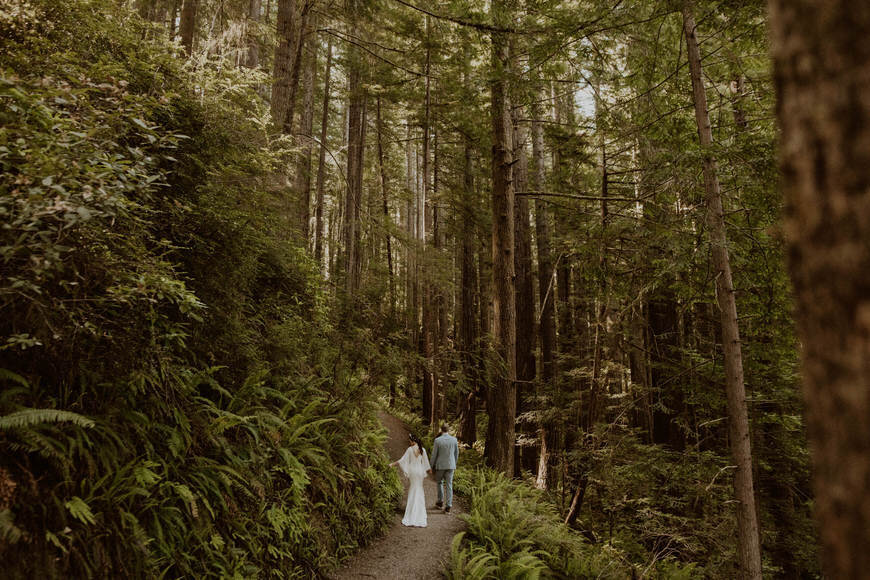 A bride and groom walking down a path in the woods.