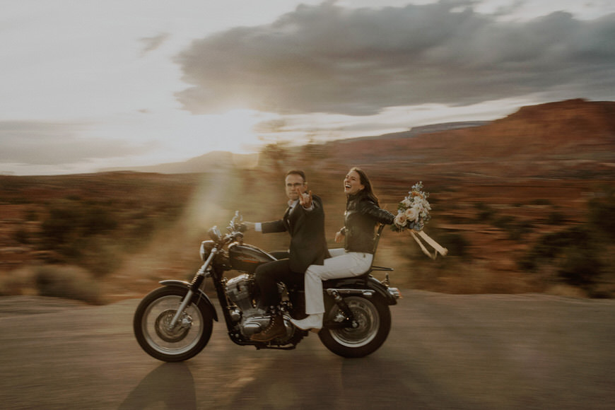 A bride and groom riding on a motorcycle in the desert.