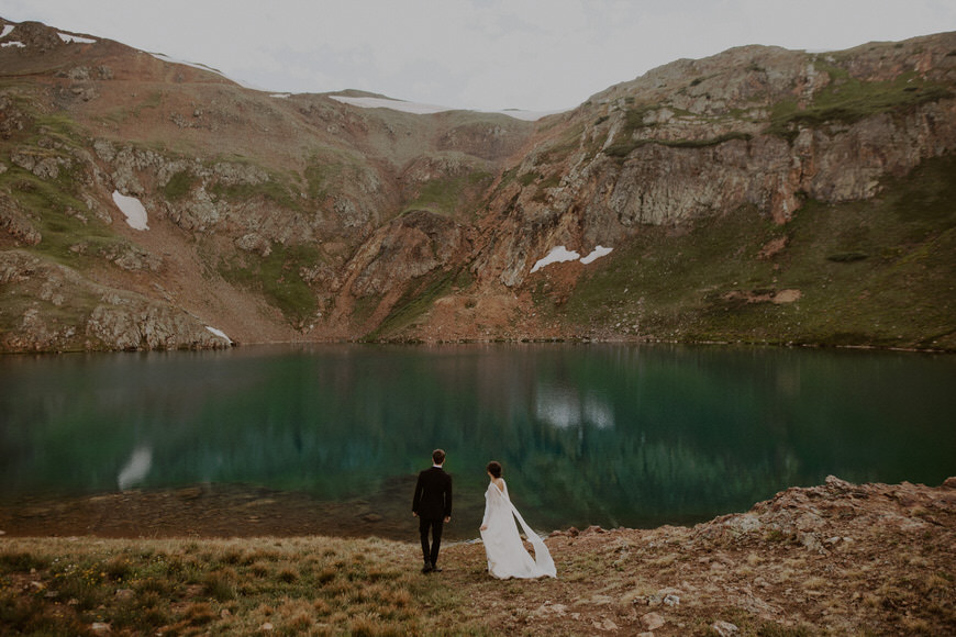 A bride and groom standing next to a lake in the mountains.