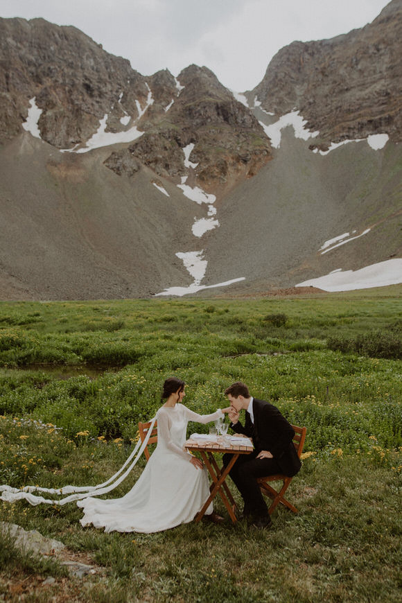 A bride and groom sit at a table in the middle of a field with mountains in the background.