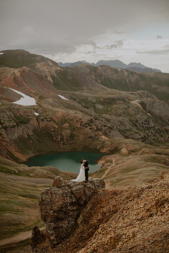 A bride and groom standing on top of a cliff overlooking a lake.