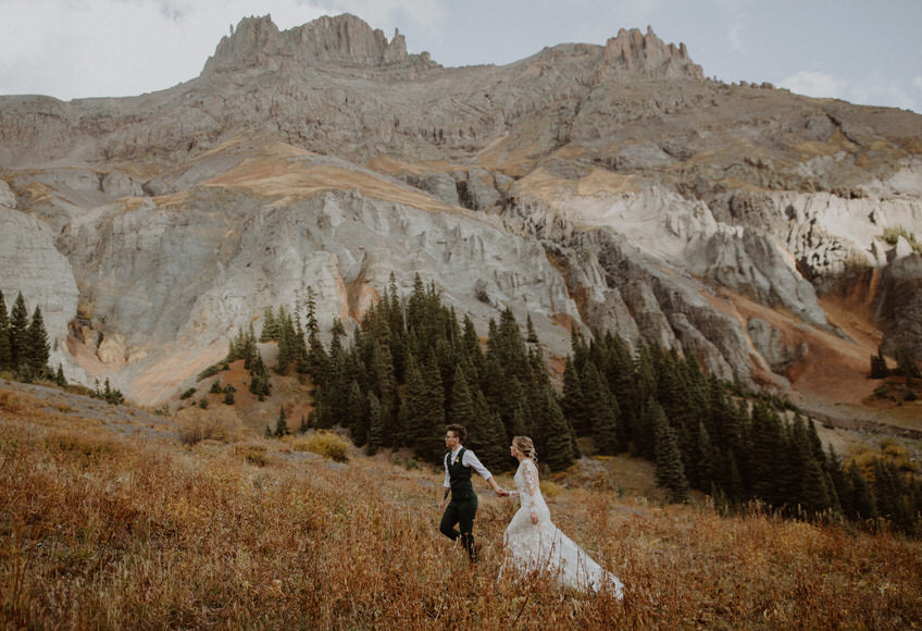 A bride and groom walking through a field with mountains in the background.