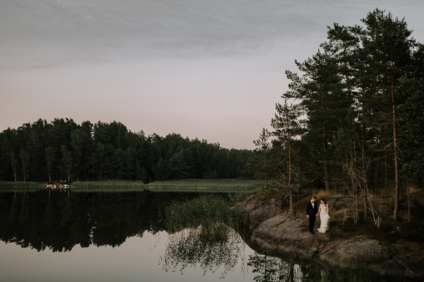A bride and groom standing on the shore of a lake.