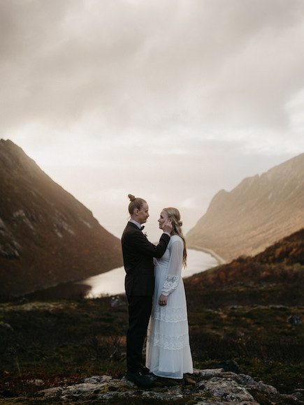 A bride and groom standing on top of a mountain overlooking a lake.