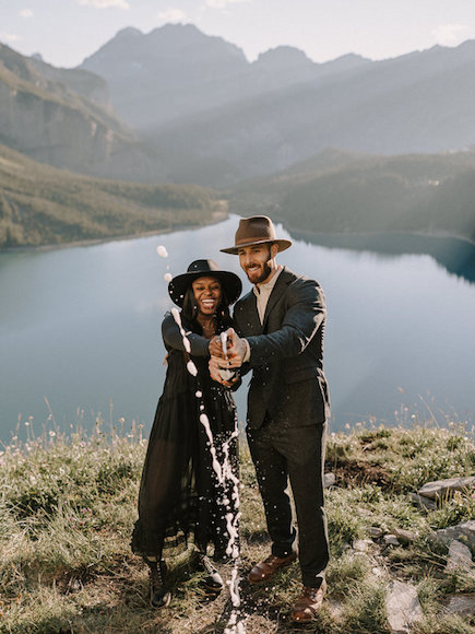 A couple in a hat and tuxedo standing in front of a lake.