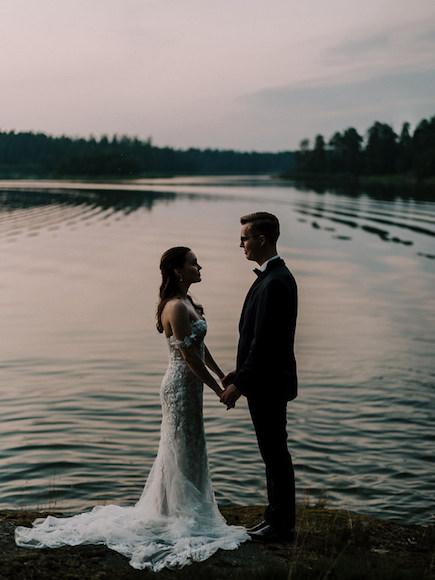 A bride and groom standing next to a lake at sunset.
