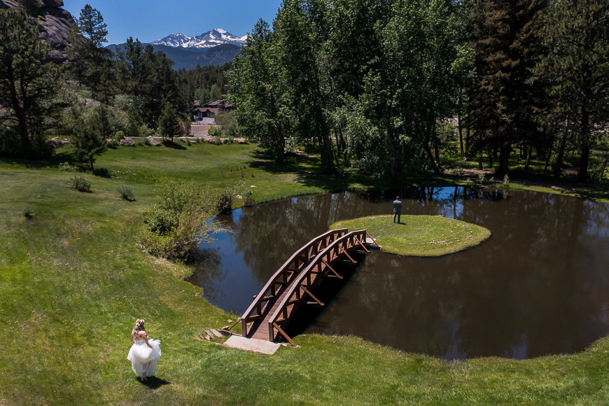 A bride walks across a bridge in front of a pond.