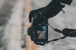 A person holding a camera on a snowy road wearing gloves.