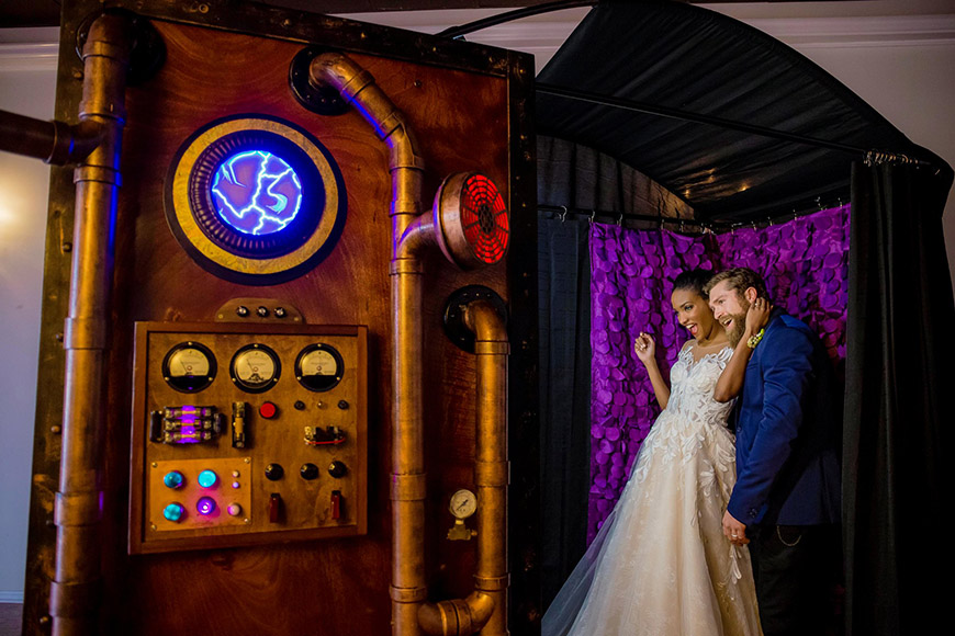 A bride and groom pose in front of a steampunk photo booth.