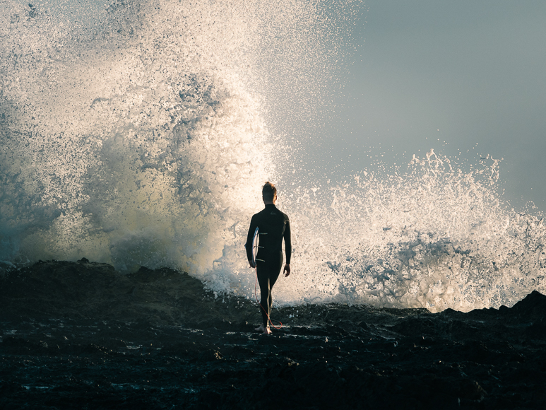 A surfer is standing on a rock in front of a large wave.